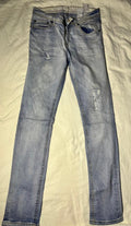 Light wash blue denim jeans with distressed rips on the thighs and knees, laid flat on a cream-colored fabric background.