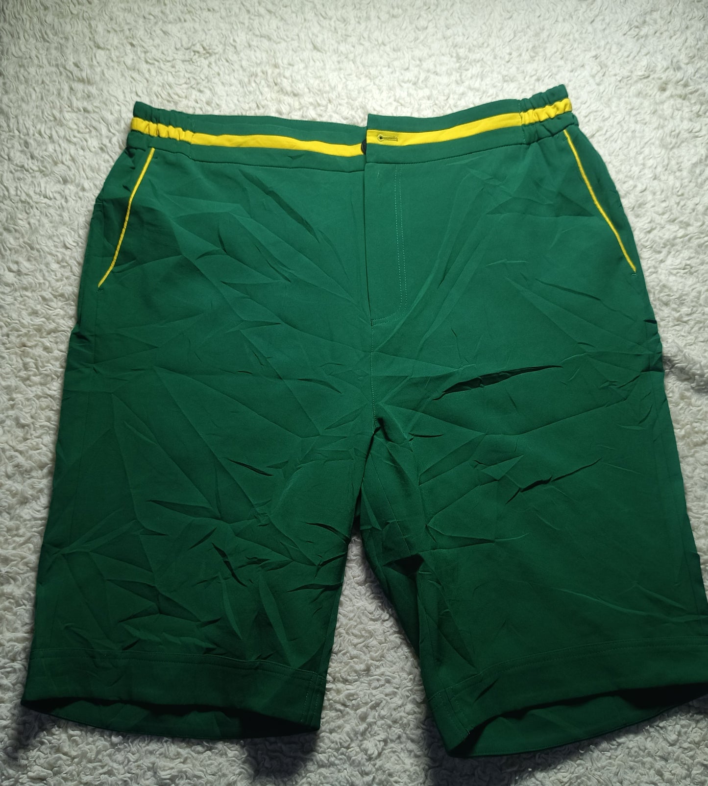 Green athletic shorts with yellow piping and a yellow elastic waistband, laid flat on a white textured rug.