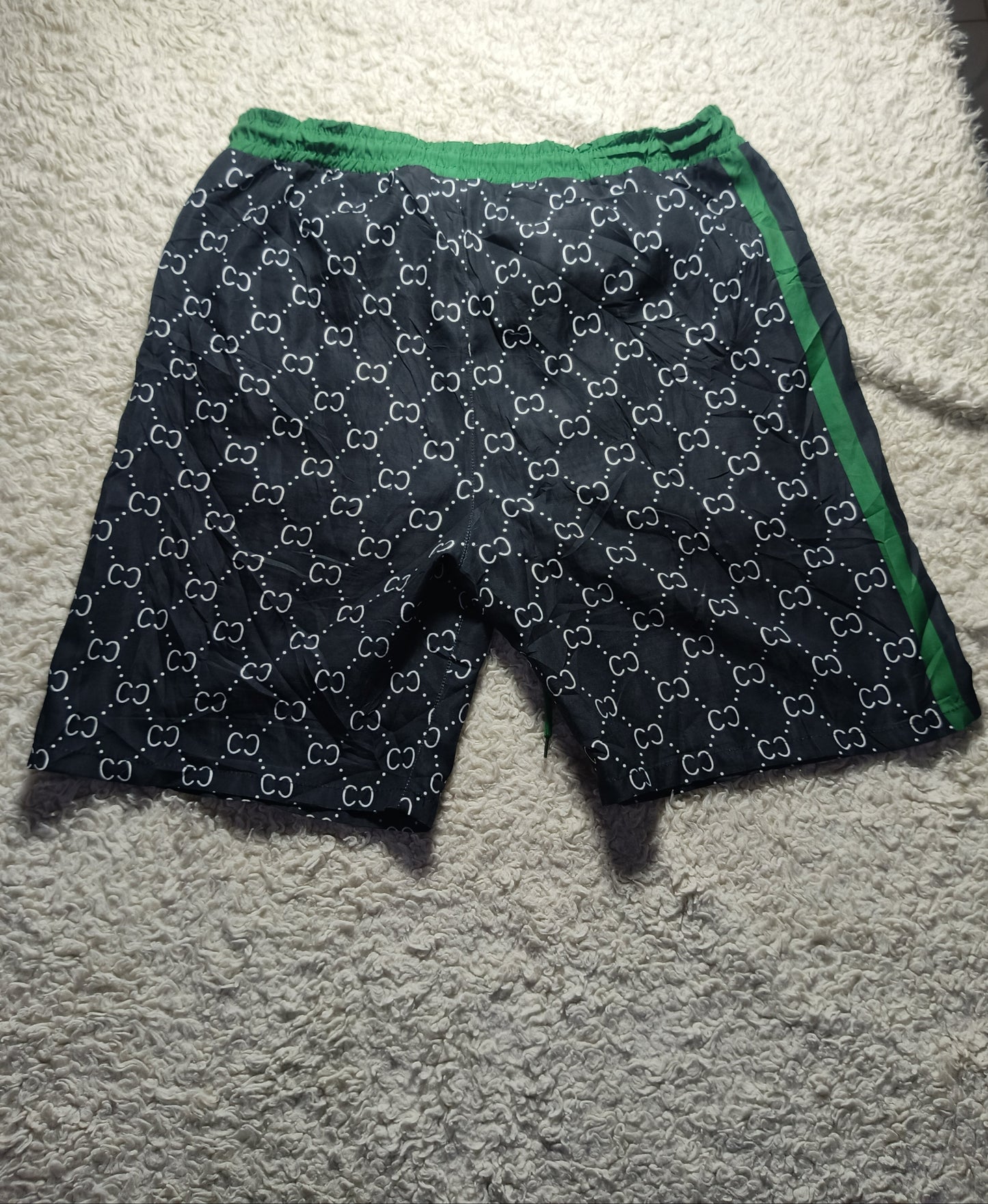 A pair of dark navy shorts with a white interlocking monogram pattern and green trim, laid flat on a textured white carpet.