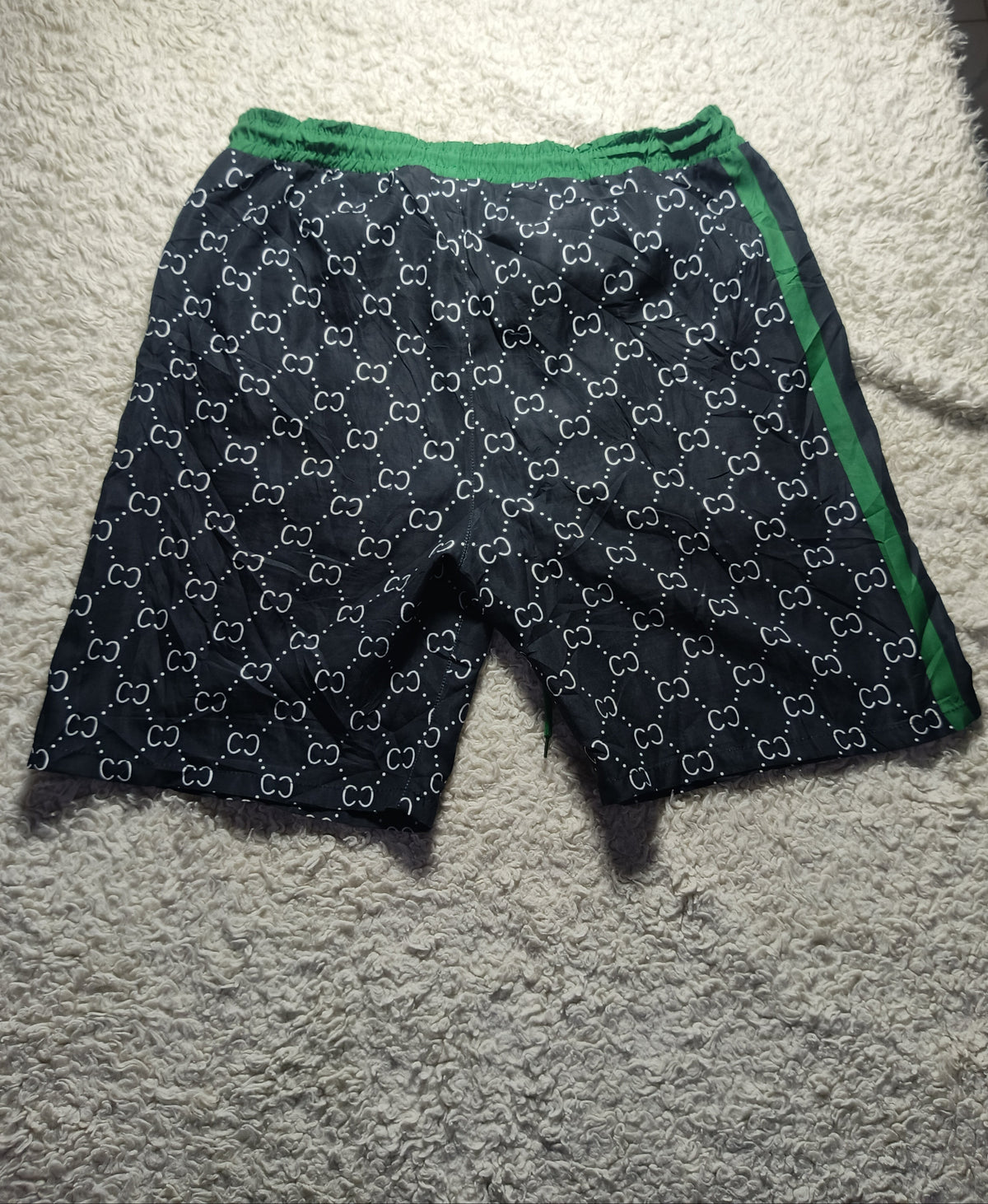 A pair of dark navy shorts with a white interlocking monogram pattern and green trim, laid flat on a textured white carpet.