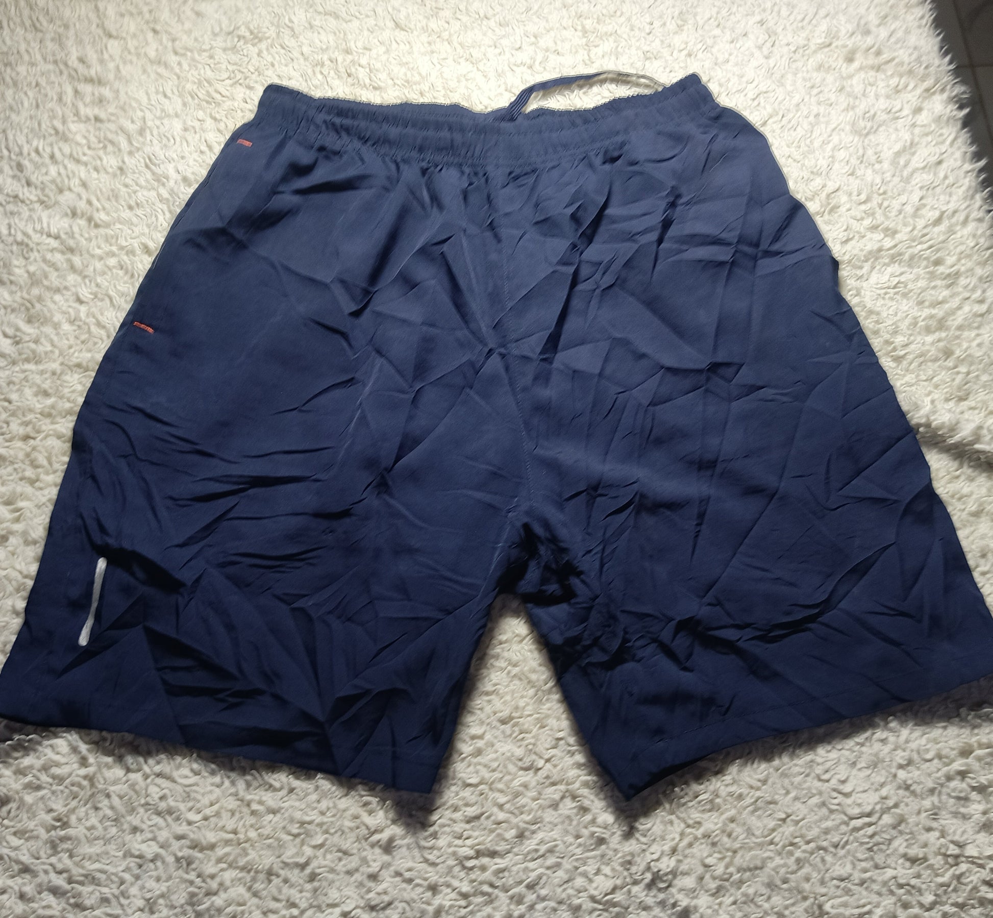 A pair of navy blue athletic shorts laid flat on a white shag rug, showing a wrinkled fabric texture and a small red logo on the upper left leg.