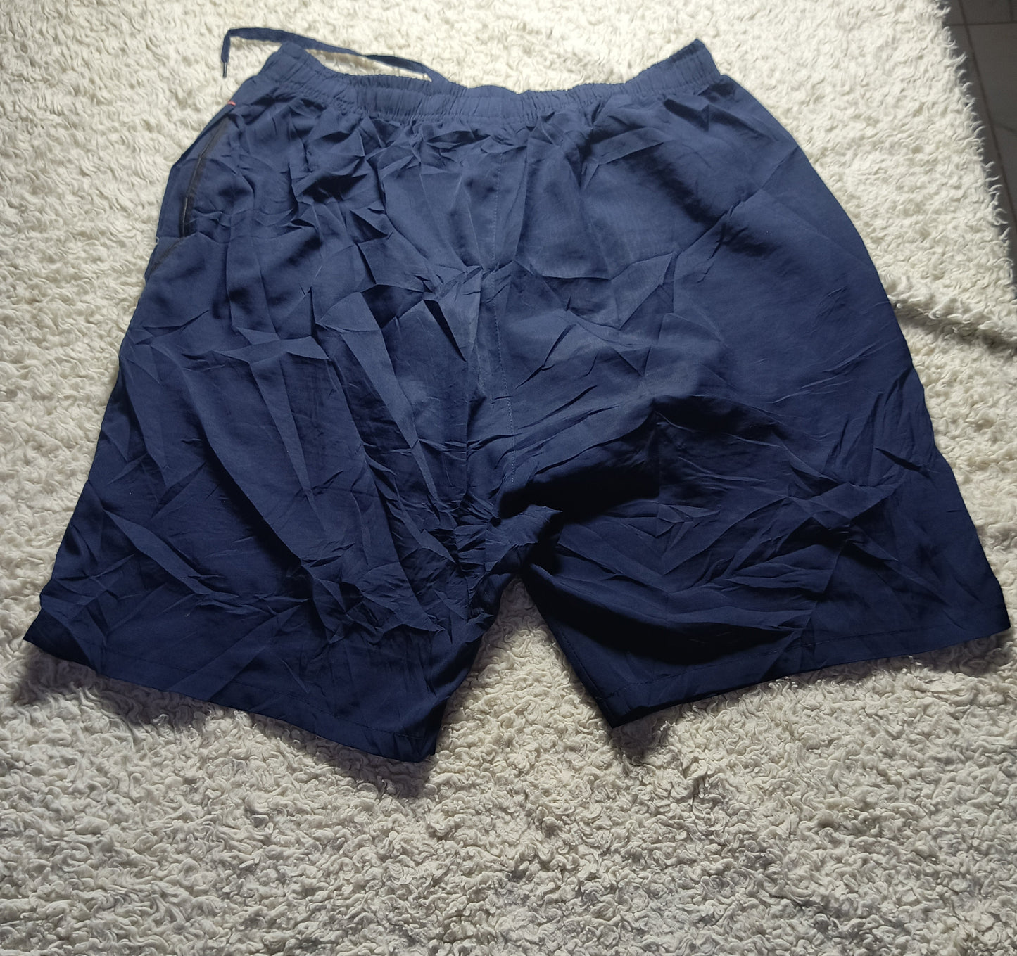 A pair of dark navy blue athletic shorts with a crinkled texture, laid flat on a light gray shag carpet. The photo is taken with a smartphone, indicated by the POVA Neo camera overlay at the bottom.