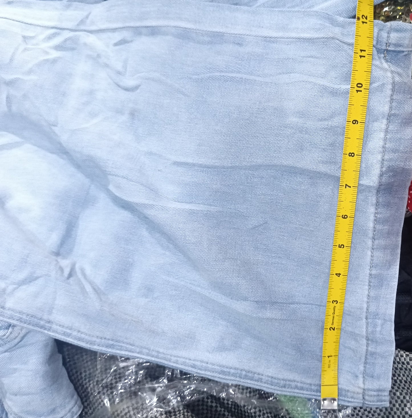 A light blue denim garment laid flat on top of other clothing, with a yellow measuring tape running vertically along the edge. A red patterned fabric and dark textured fabric are visible around it, and a white text strip appears at the bottom with camera metadata.