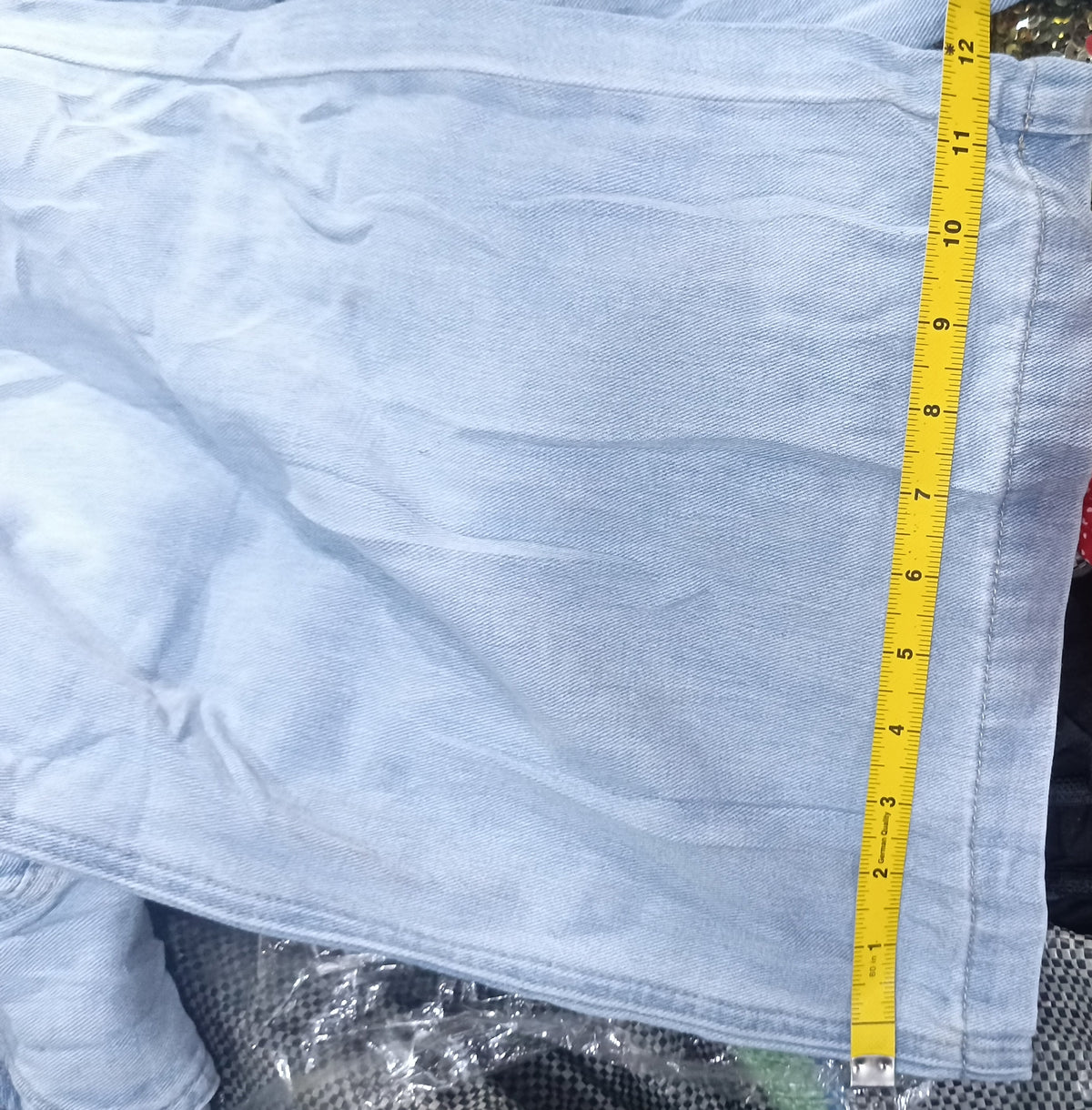 A light blue denim garment laid flat on top of other clothing, with a yellow measuring tape running vertically along the edge. A red patterned fabric and dark textured fabric are visible around it, and a white text strip appears at the bottom with camera metadata.