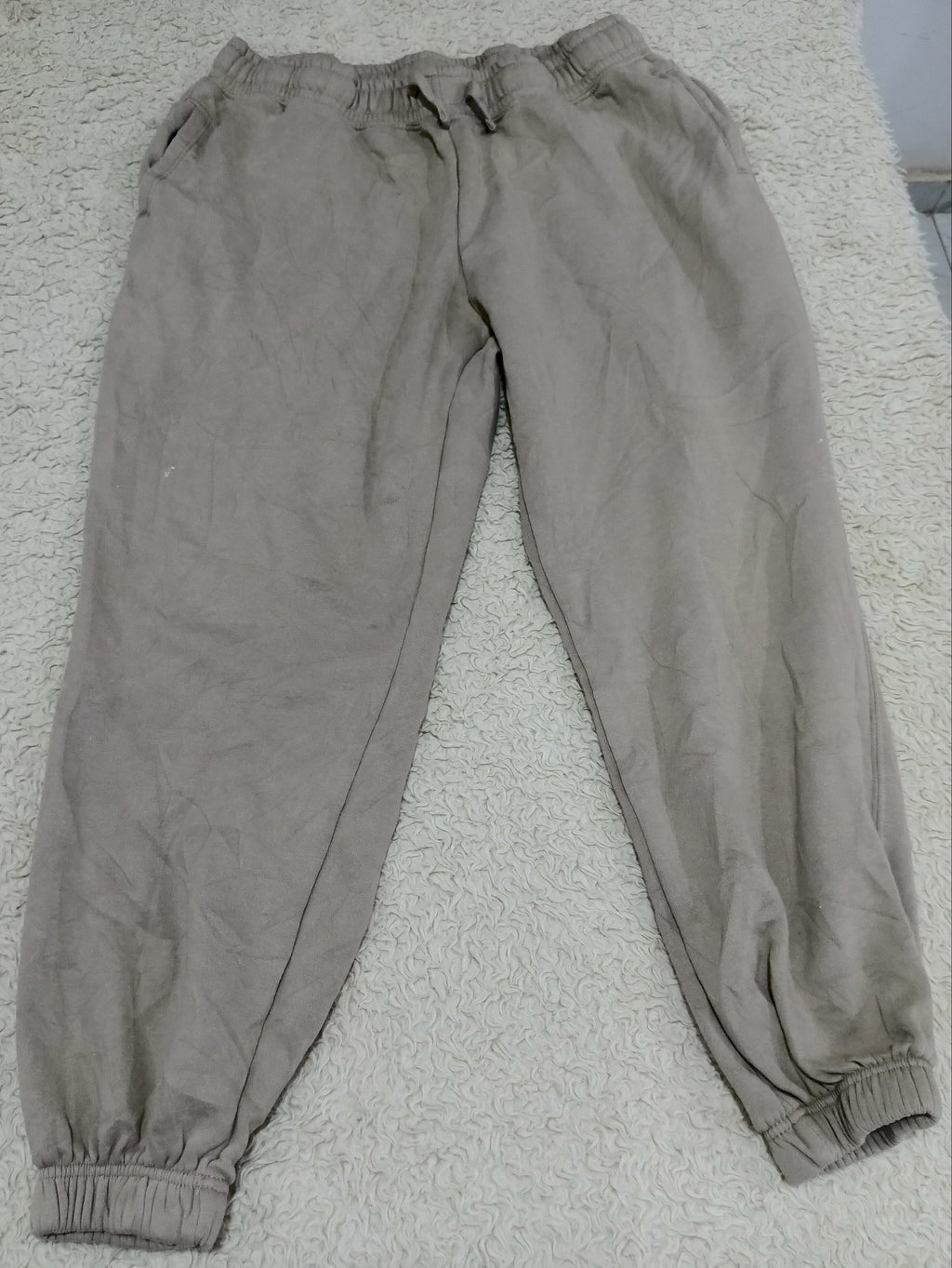 A pair of light brown or taupe jogger pants laid flat on a textured white carpet. The pants feature an elastic waistband with a drawstring, side pockets, and elastic cuffs at the ankles.