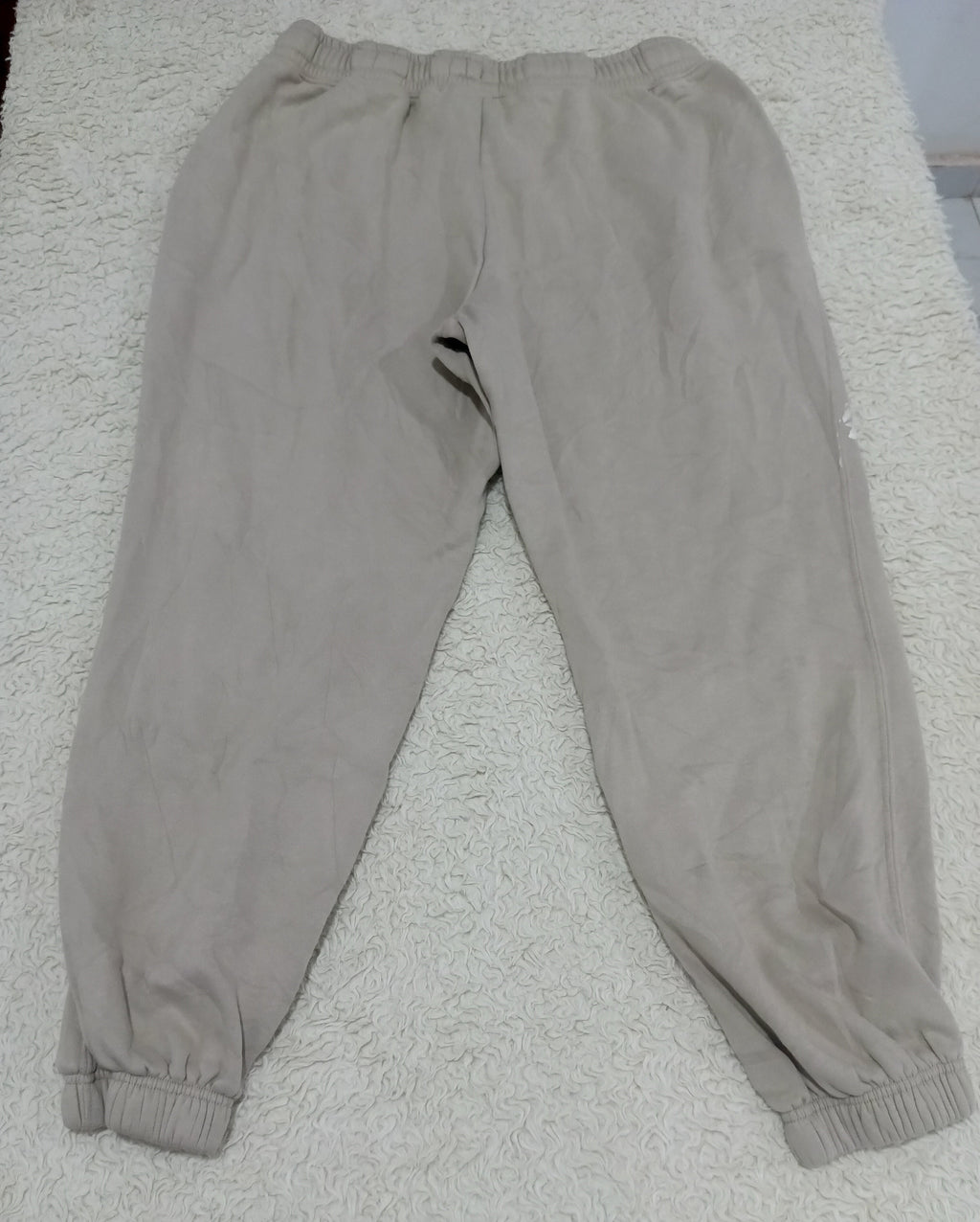 A pair of light beige jogger sweatpants with elastic waistbands and ribbed ankle cuffs, laid flat on a white textured rug.