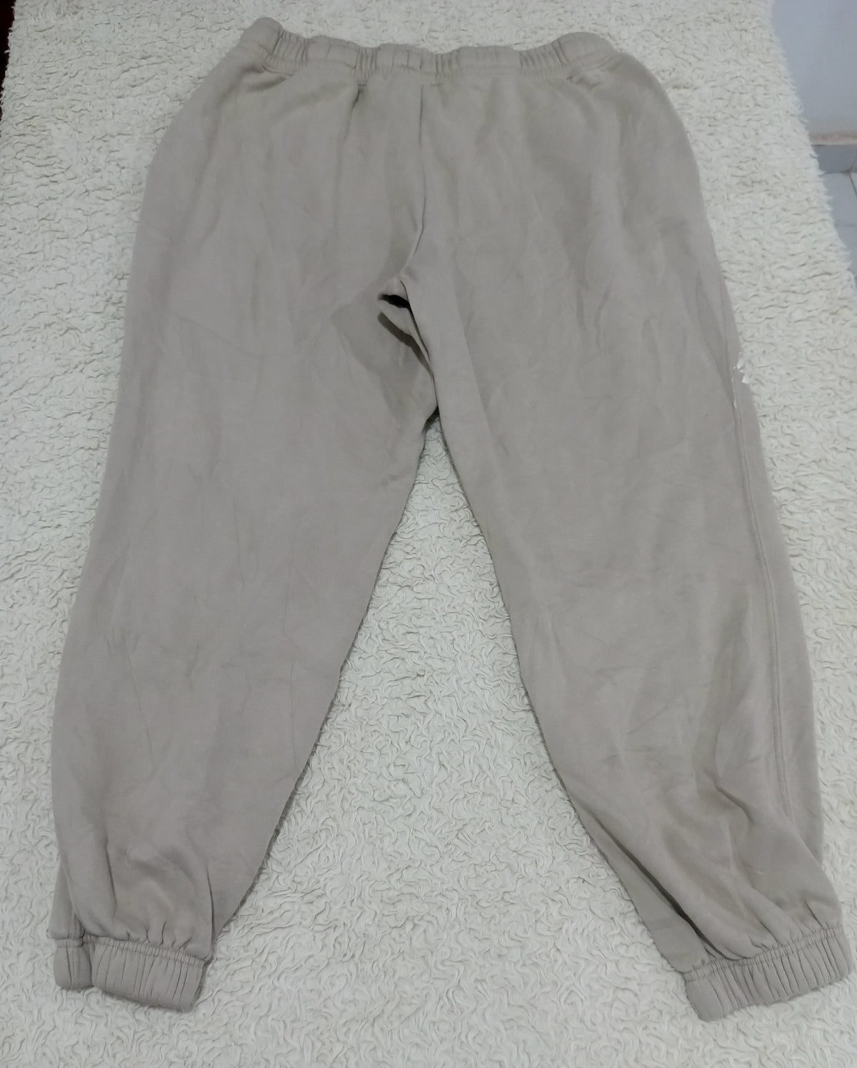 A pair of light beige jogger sweatpants with elastic waistbands and ribbed ankle cuffs, laid flat on a white textured rug.