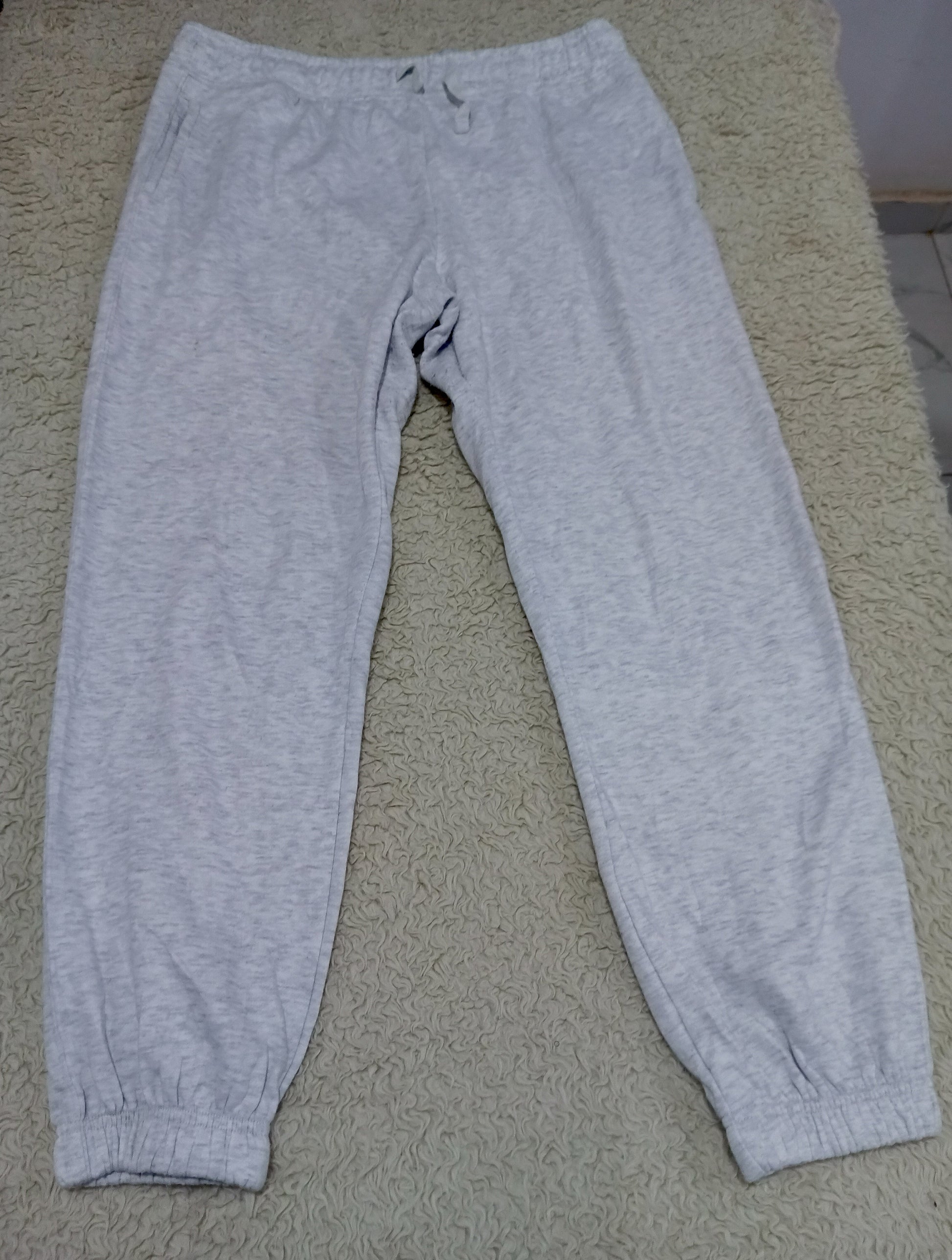 Light gray heather sweatpants with an elastic waistband and cuffed ankles, laid flat on a beige textured carpet.