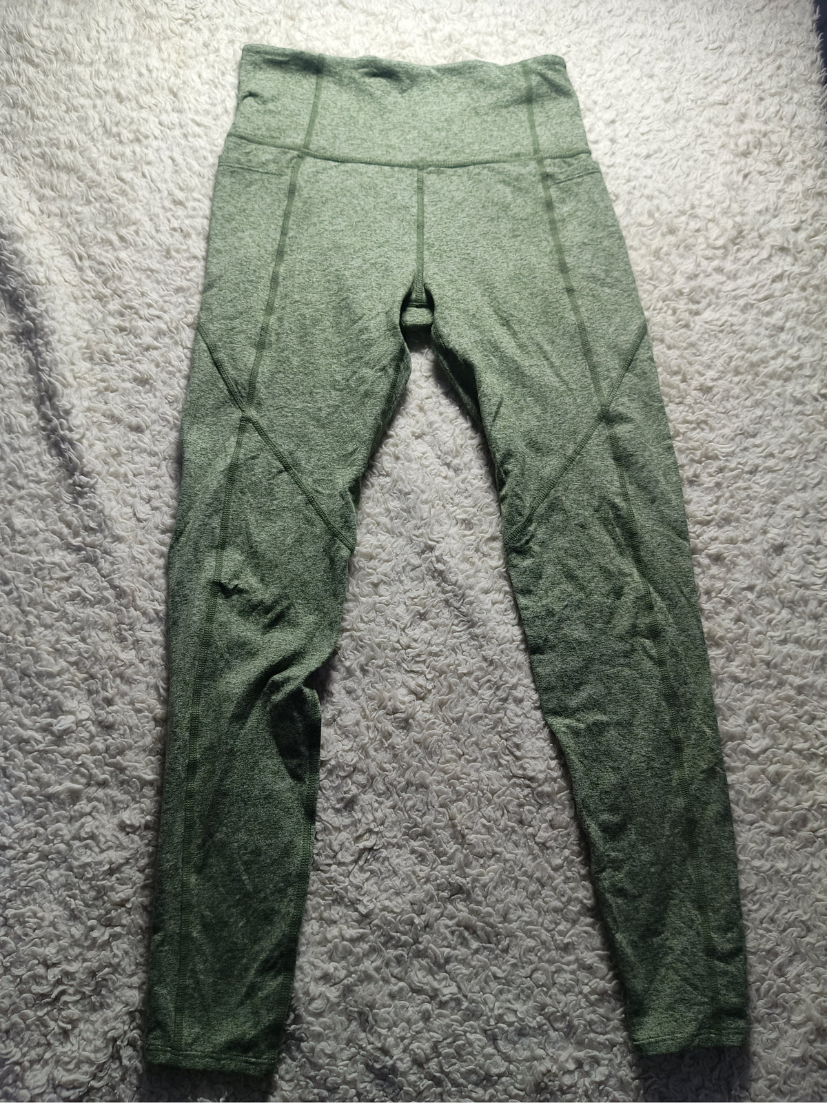 A pair of high-waisted, form-fitting leggings in a heathered sage green color, laid flat on a textured gray carpet.