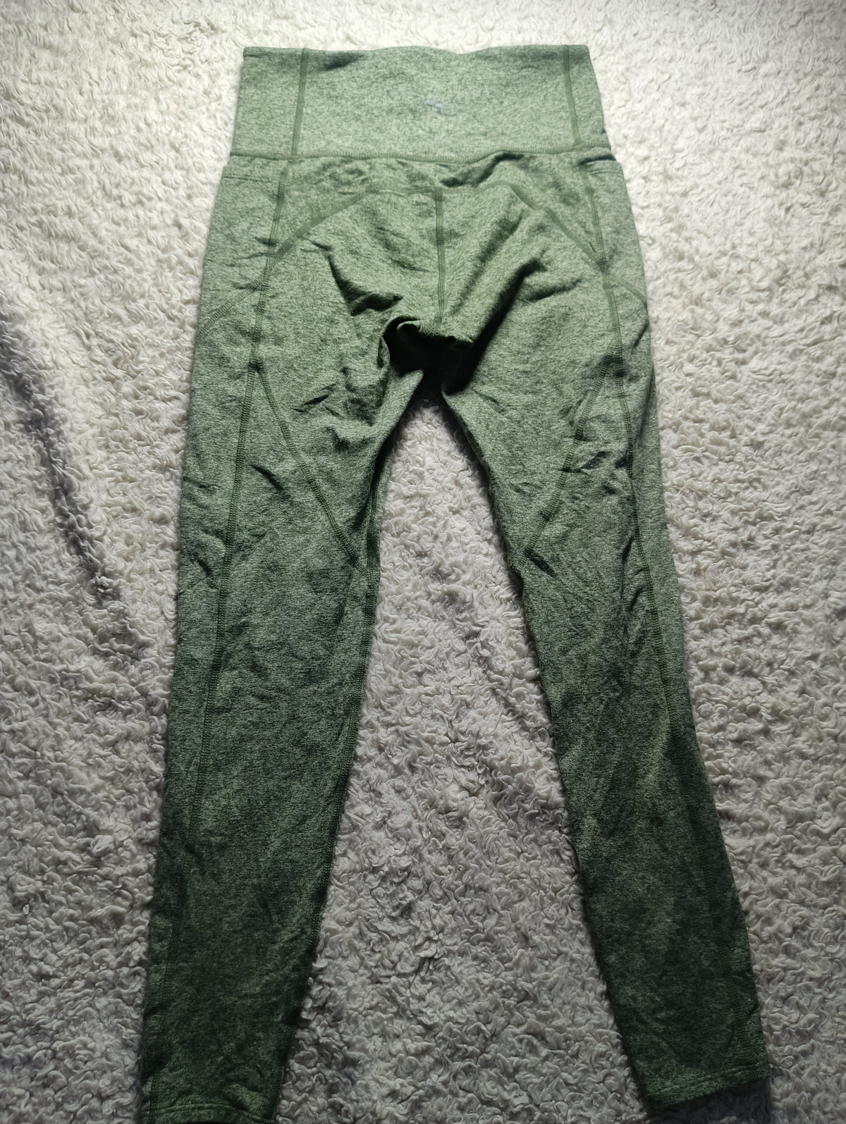 A pair of olive green high-waisted leggings laid flat on a textured gray carpet. The image has a white border at the bottom with camera metadata 'POVA & Neo 23mm f/1.