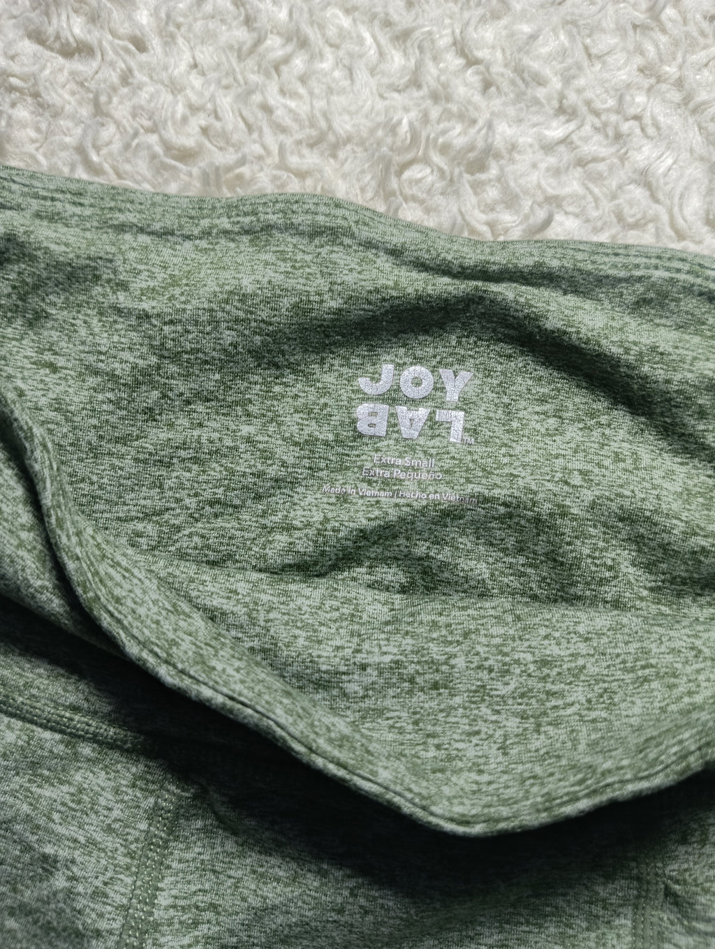 A close-up of a folded heathered sage green garment, likely a hoodie or sweatshirt, with a white printed 'JOY LAB' logo on the chest. The fabric is laid flat on a textured white surface.