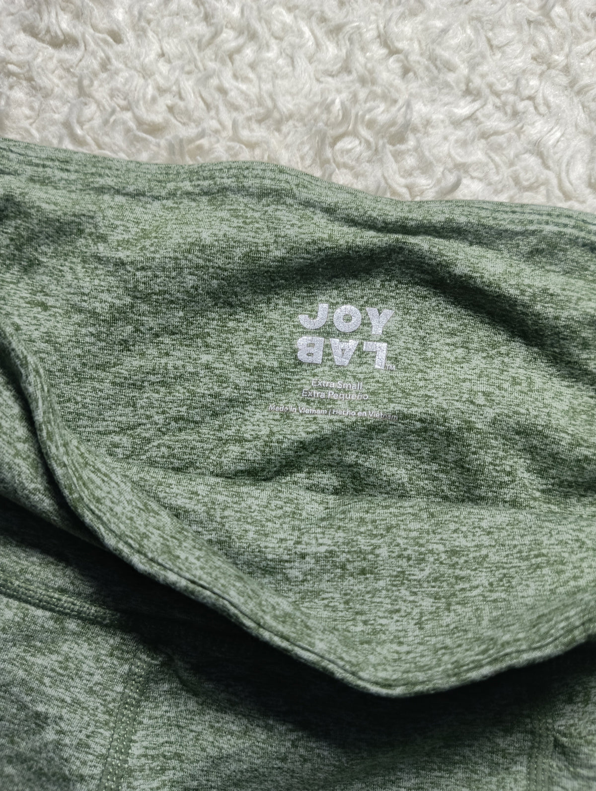 A close-up of a folded heathered sage green garment, likely a hoodie or sweatshirt, with a white printed 'JOY LAB' logo on the chest. The fabric is laid flat on a textured white surface.