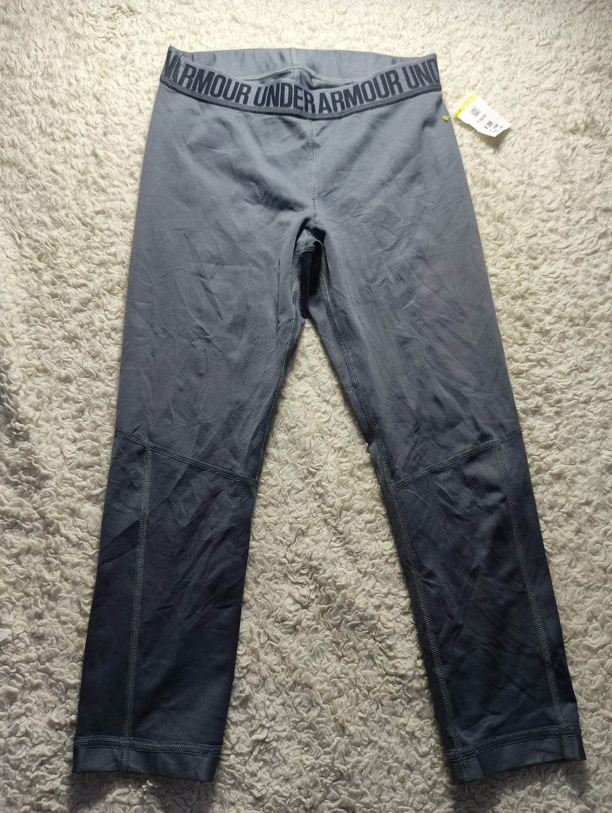 A pair of dark gray Under Armour gradient leggings laid flat on a beige carpet. The waistband features the Under Armour logo in white, and a small white product tag is attached near the right hip.