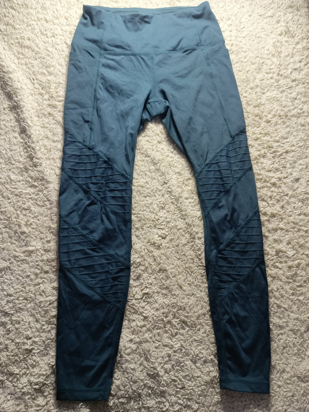 A pair of dark teal athletic leggings laid flat on a textured beige carpet. The leggings feature a smooth upper body and textured, ribbed knee panels.