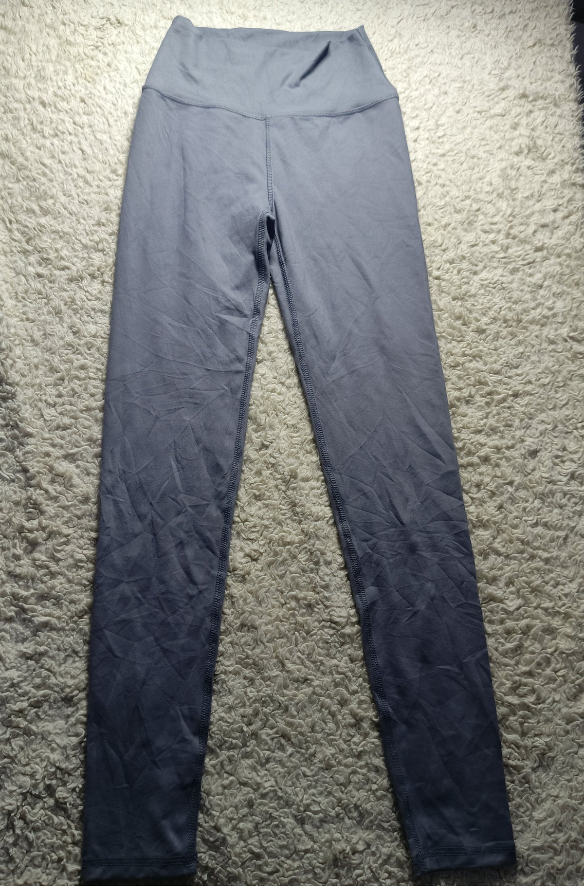A pair of high-waisted gray leggings laid flat on a textured beige carpet. The leggings have a subtle gradient effect, fading from a medium gray at the top to a very dark charcoal gray at the bottom.