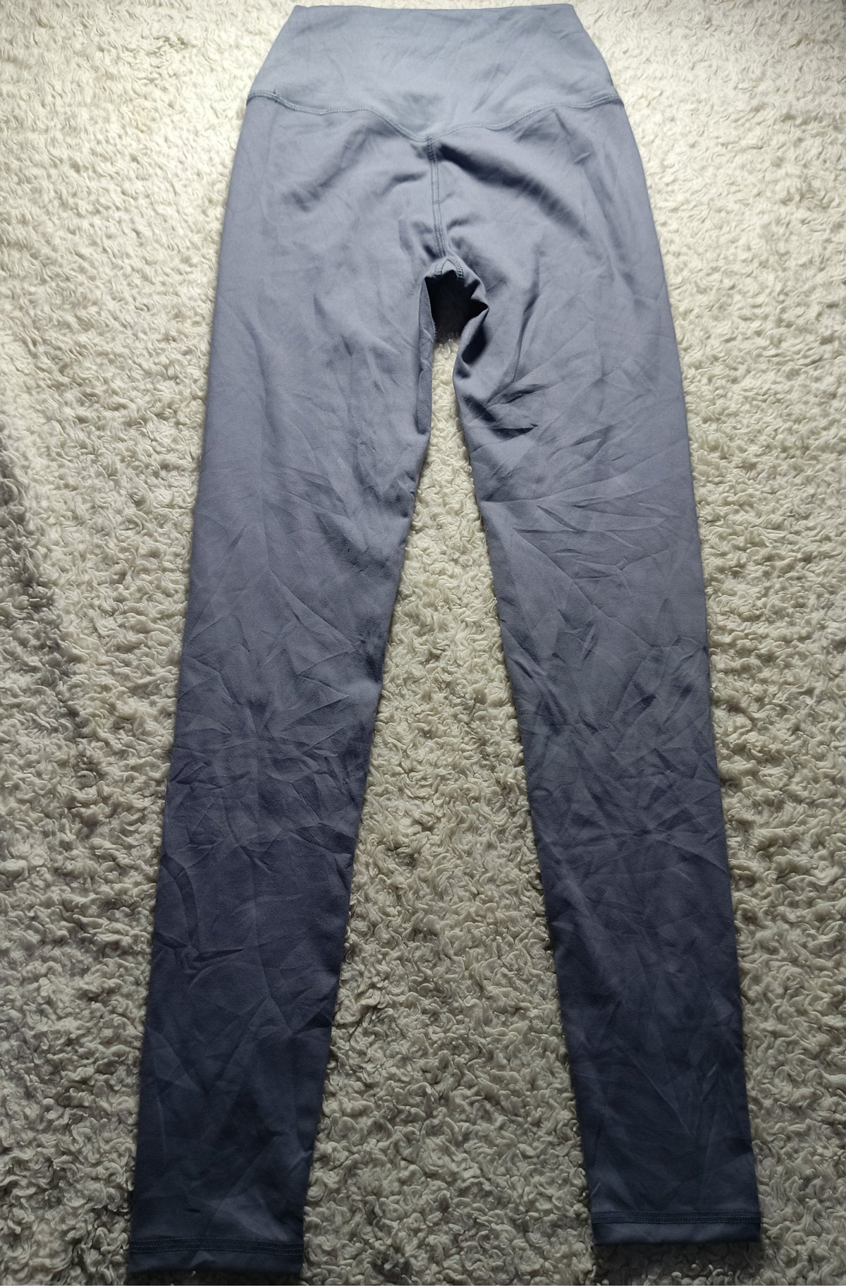 A pair of dark gray high-waisted leggings laid flat on a textured beige rug. The image has a camera watermark at the bottom indicating 'POVA 6 Neo' and camera settings.