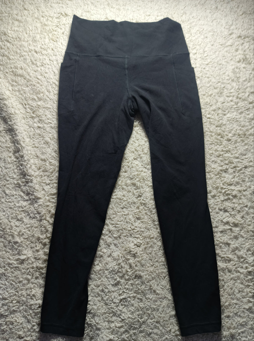 Black leggings laid flat on a textured beige carpet, photographed from above with a camera watermark at the bottom.