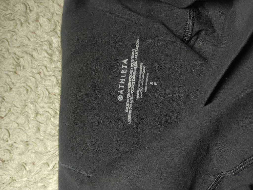 Close-up of a black athletic garment showing the inner neck label with the brand name ATHLETA and size information, photographed on a textured cream surface.
