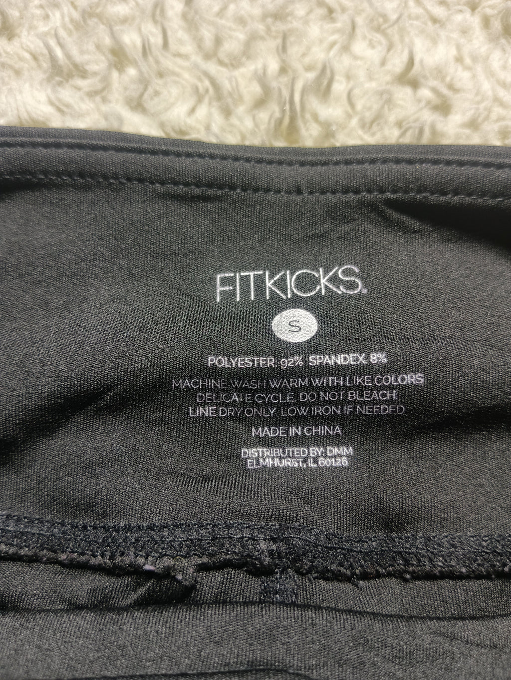 Close-up of the inside neck label of a black Fitkicks garment, showing size S details and care instructions, photographed against a textured cream background.