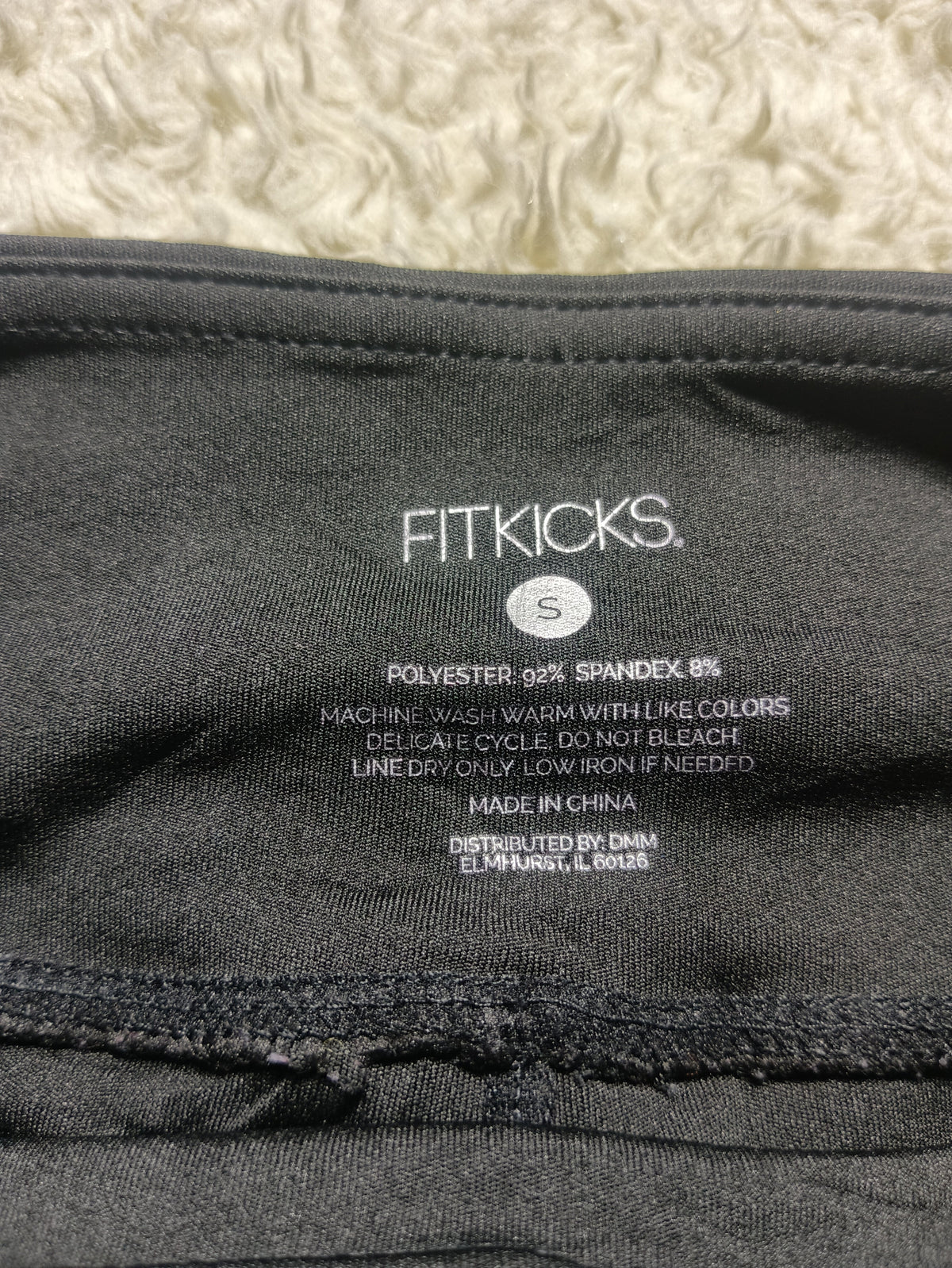 Close-up of the inside neck label of a black Fitkicks garment, showing size S details and care instructions, photographed against a textured cream background.