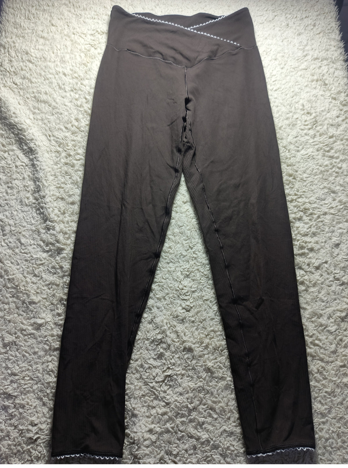 A pair of dark brown high-waisted leggings laid flat on a textured gray carpet. The leggings feature a crossover waistband with white contrast stitching and a small white embroidered detail on the left hip.