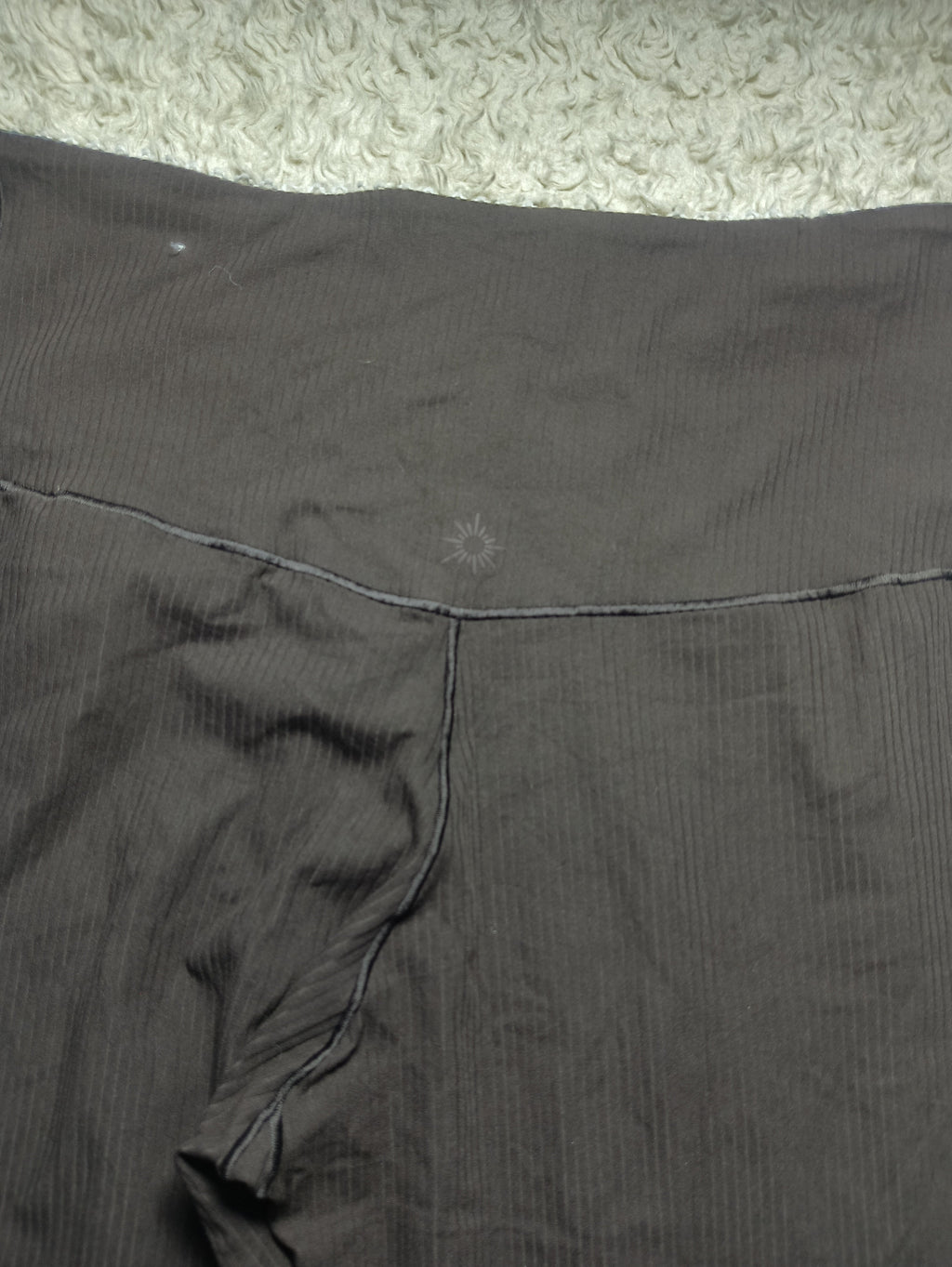 Close-up of dark gray ribbed fabric, likely a garment or textile panel, laid flat on a light textured surface. A faint embroidered logo is visible near the center, and camera metadata appears along the bottom.