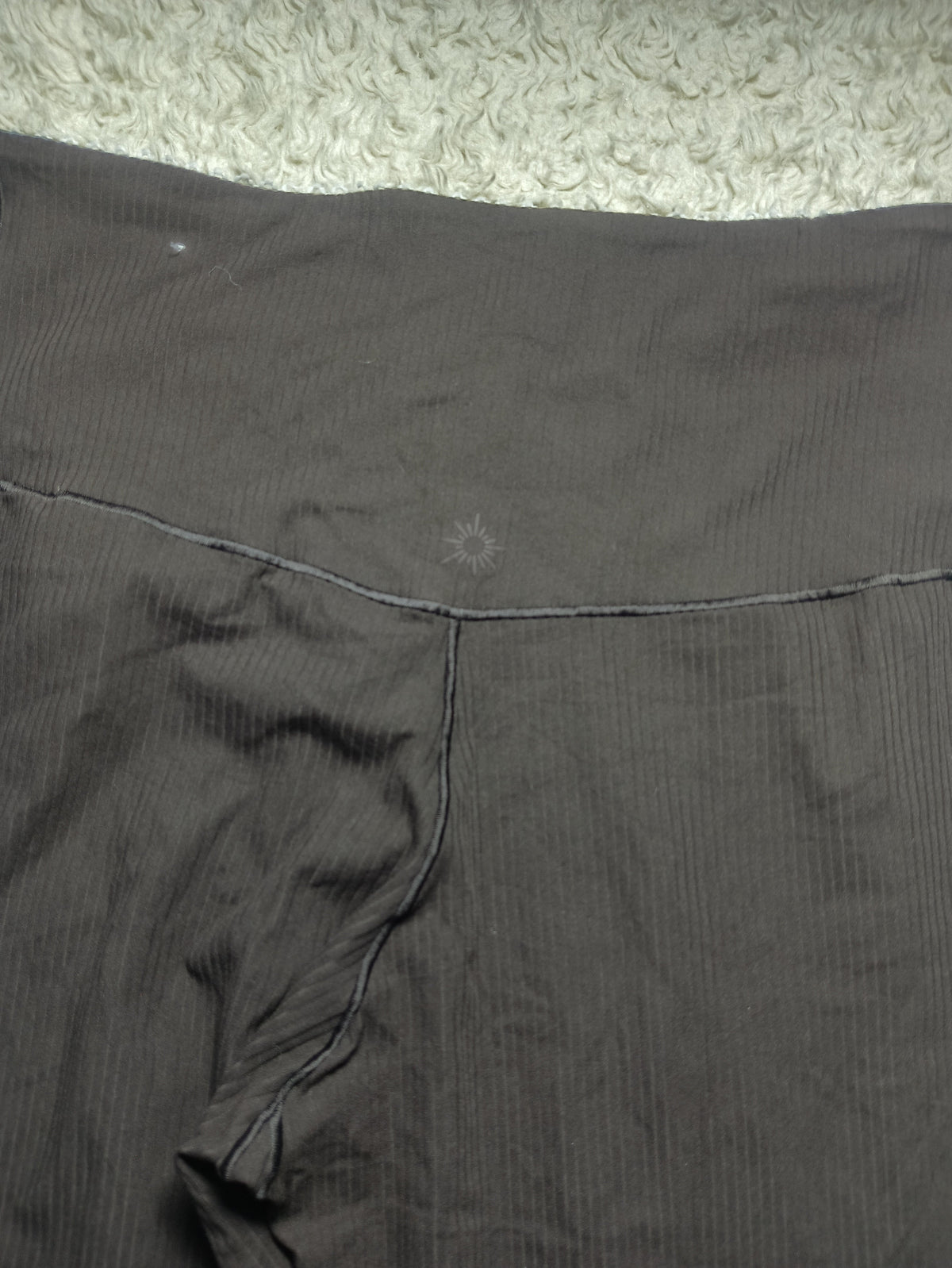 Close-up of dark gray ribbed fabric, likely a garment or textile panel, laid flat on a light textured surface. A faint embroidered logo is visible near the center, and camera metadata appears along the bottom.