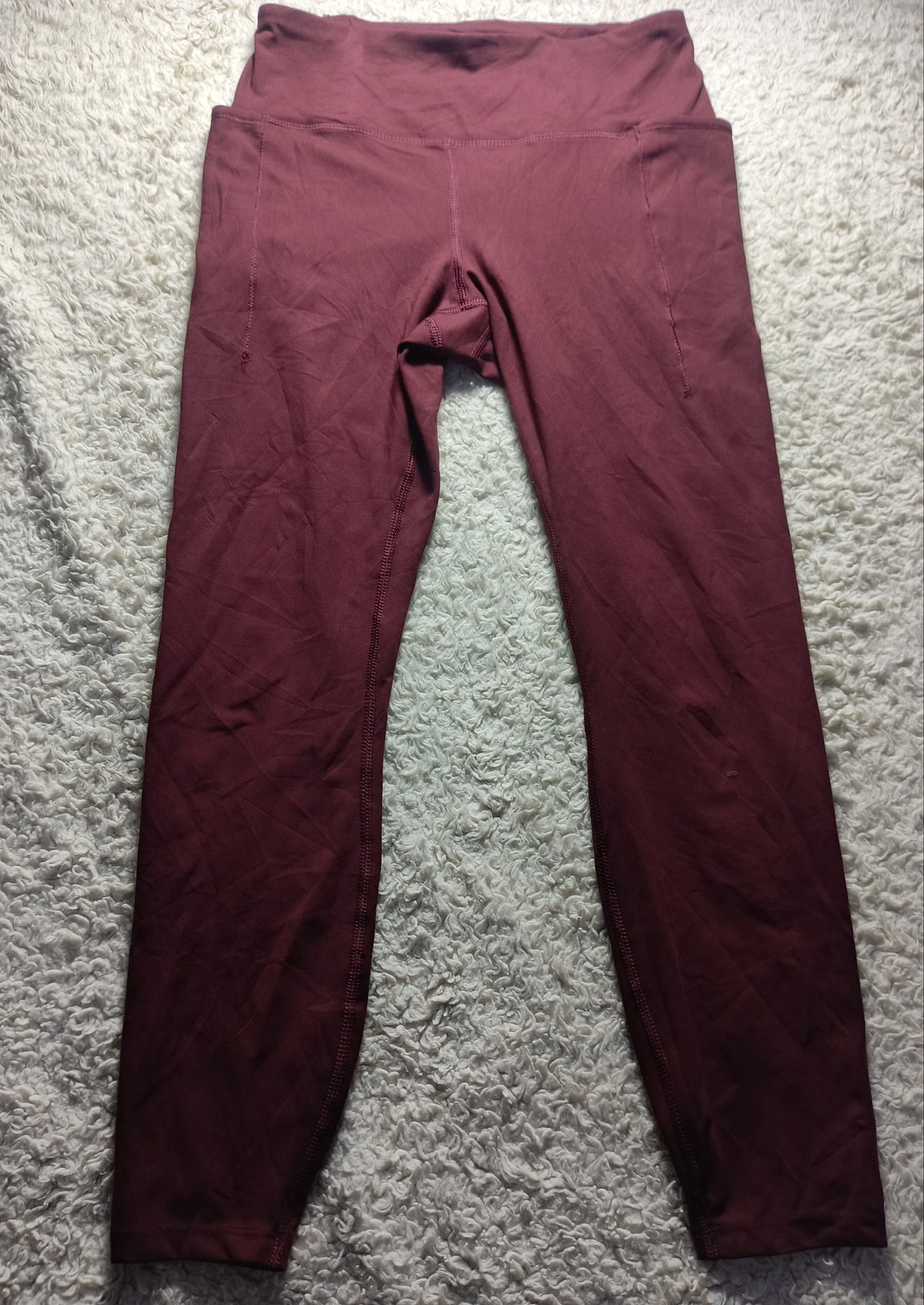 A pair of high-waisted leggings in a deep burgundy or maroon color, laid flat on a textured gray carpet. The image has a camera watermark at the bottom.