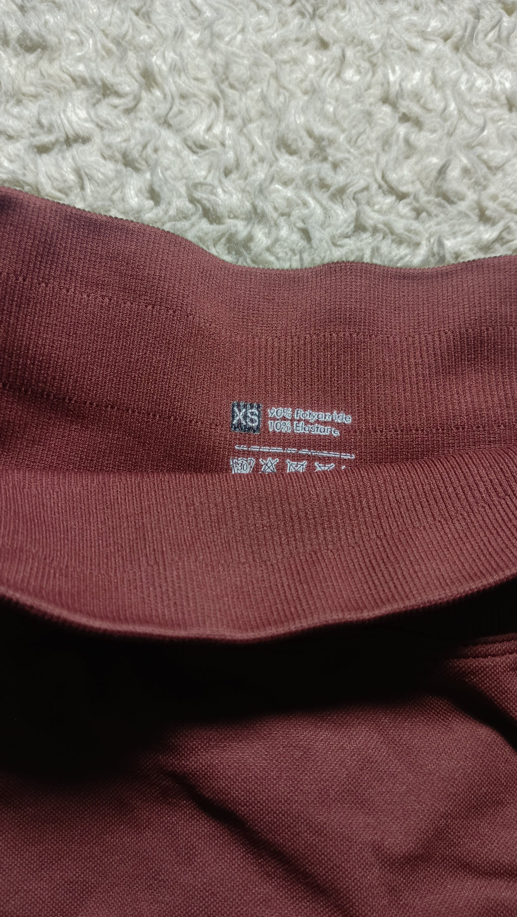 Close-up of a burgundy ribbed knit garment, likely a sweatshirt or hoodie, showing the inner collar with size and care labels. The garment is laid flat on a textured white rug.