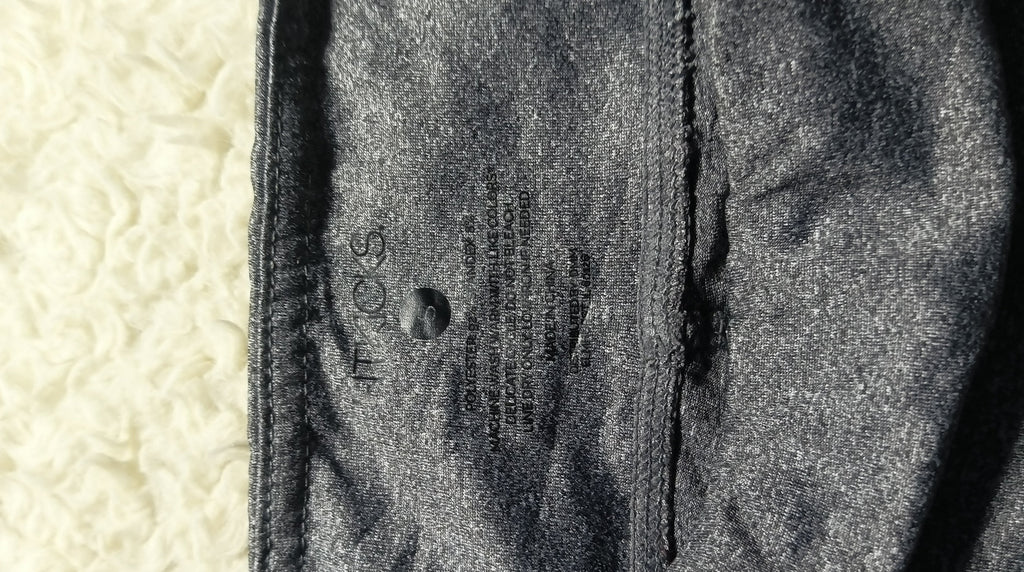 Close-up of a dark gray textured fabric garment with a visible seam and printed care label, placed against a light cream textured background. Camera metadata text is visible at the bottom.