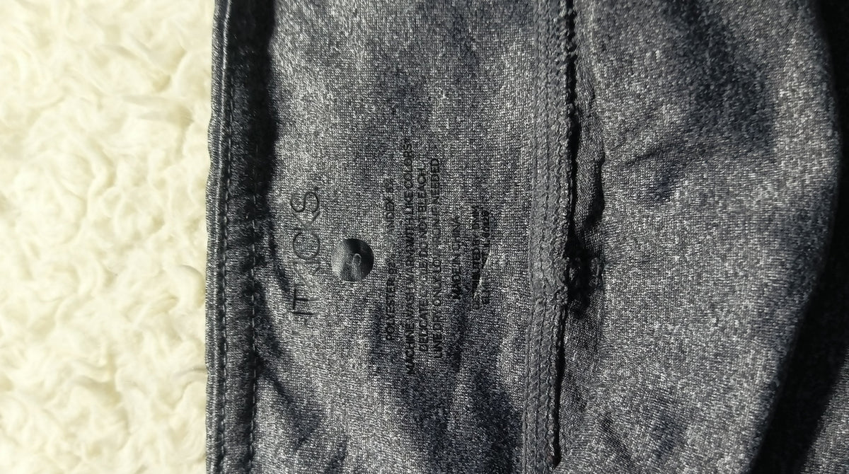Close-up of a dark gray textured fabric garment with a visible seam and printed care label, placed against a light cream textured background. Camera metadata text is visible at the bottom.