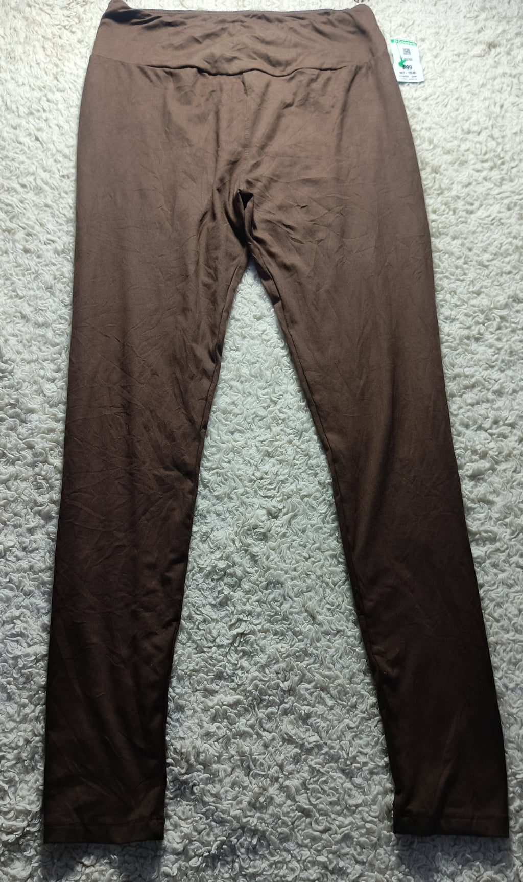 A pair of solid brown leggings laid flat on a textured gray carpet, with a small retail tag visible at the waistband.