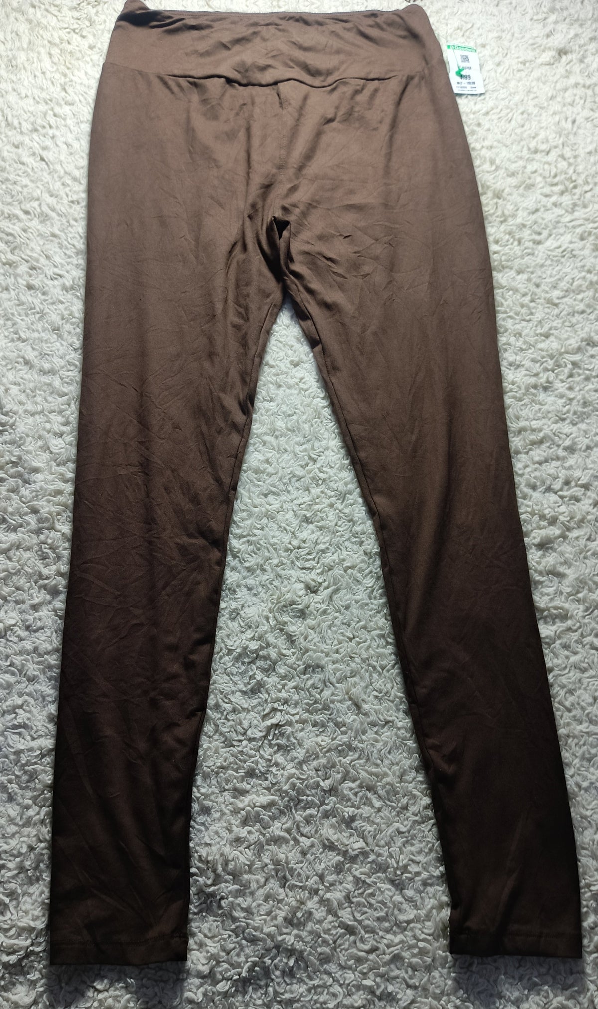 A pair of solid brown leggings laid flat on a textured gray carpet, with a small retail tag visible at the waistband.