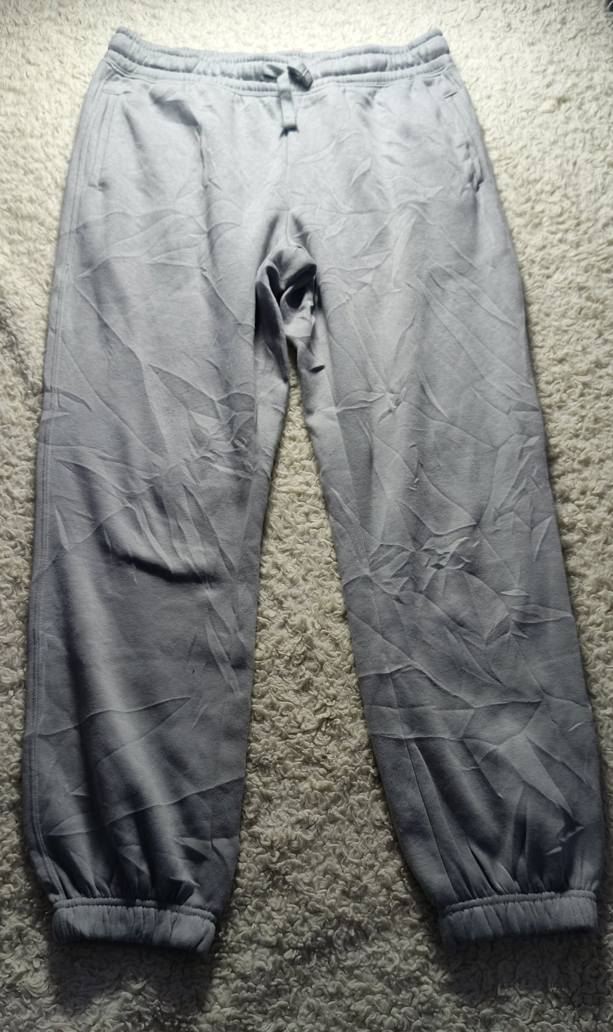 A pair of light gray jogger pants laid flat on a textured gray carpet. The pants have a drawstring waist, elastic cuffs, and a wrinkled, creased texture.