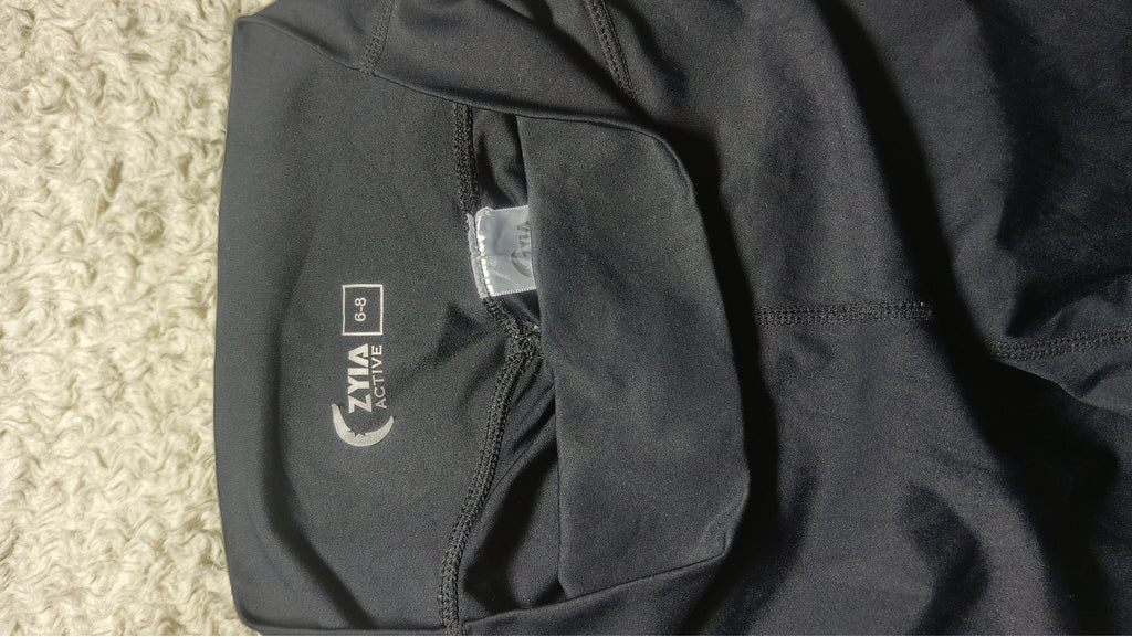 Close-up of a black athletic garment laid flat on a textured beige carpet, showing a side pocket with a visible ZIMA ACTIVE logo and a small sewn label.
