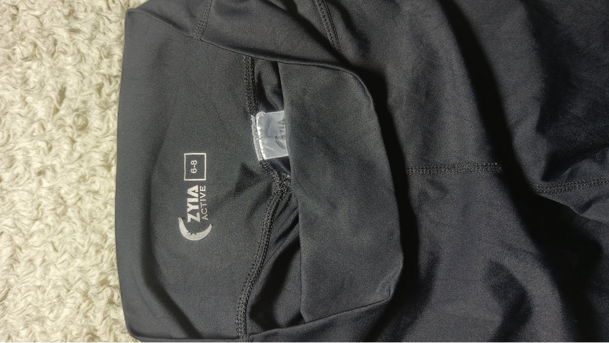 Close-up of a black athletic garment laid flat on a textured beige carpet, showing a side pocket with a visible ZIMA ACTIVE logo and a small sewn label.