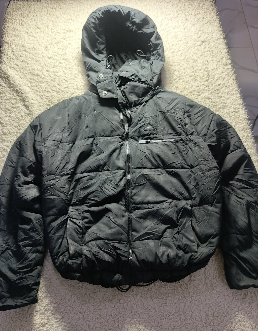A black hooded puffer jacket laid flat on a textured beige rug. The jacket has a quilted design, a front zipper, and a zippered chest pocket.