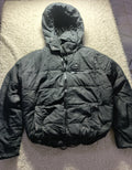 A black hooded puffer jacket laid flat on a textured beige rug. The jacket has a quilted design, a front zipper, and a zippered chest pocket.