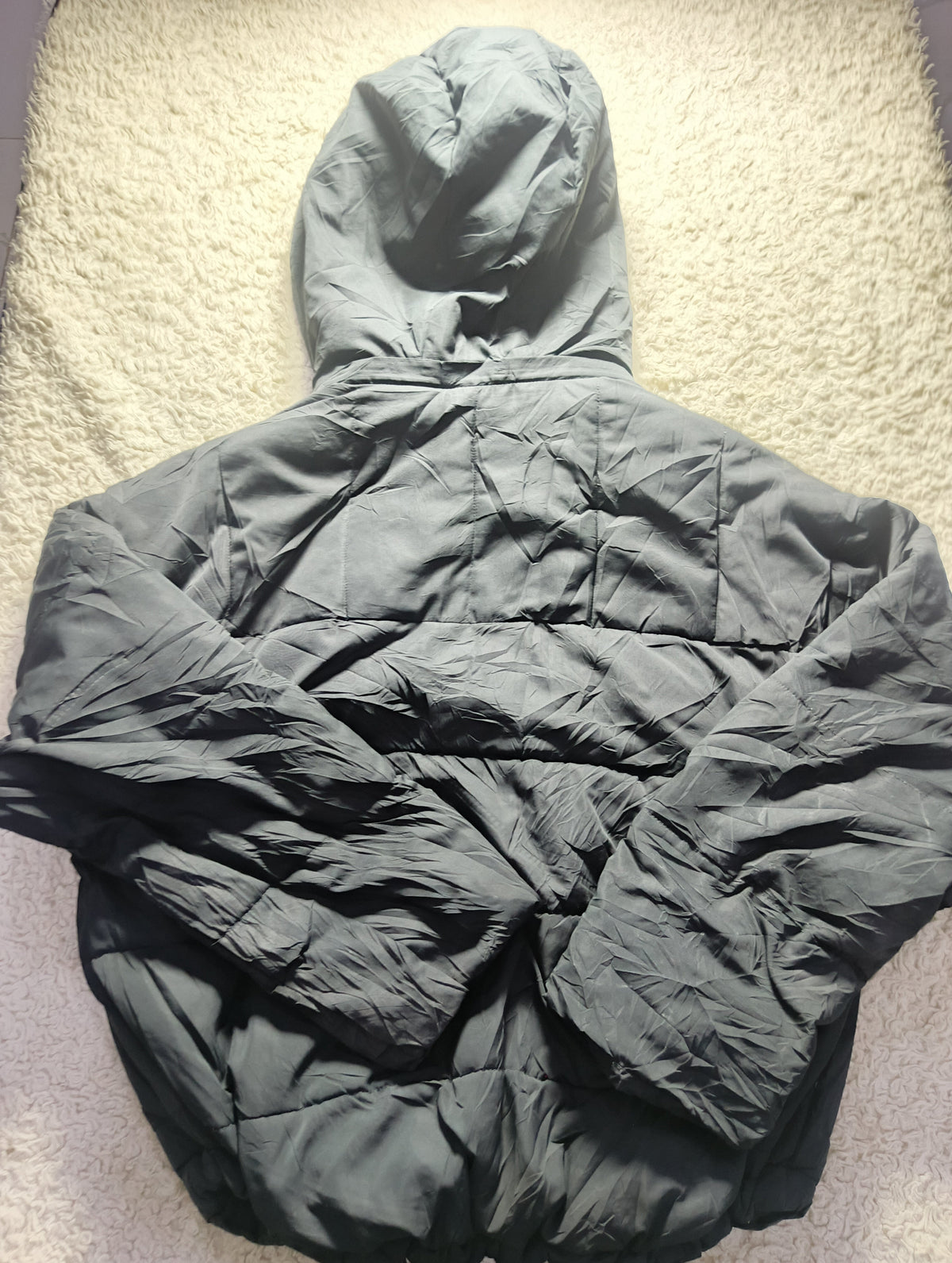 A dark gray hooded puffer jacket laid flat on a textured beige carpet. The image has a camera watermark at the bottom.
