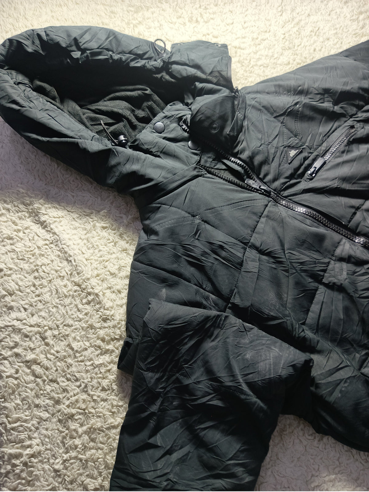 A black puffer jacket laid flat on a textured beige carpet, with a visible front zipper and quilted padding.