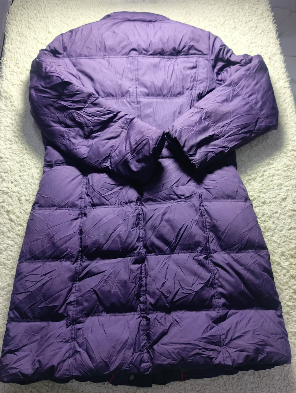 A purple quilted puffer coat laid flat on a textured white carpet, photographed from above. The image includes a camera watermark at the bottom.