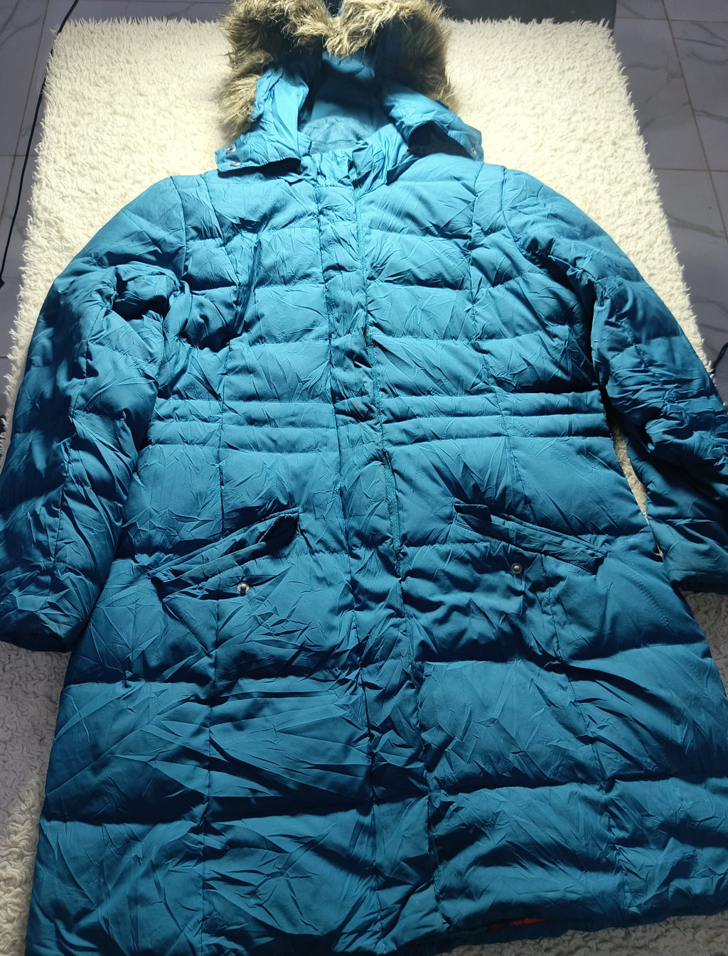 A teal blue puffer jacket with a fur-trimmed hood, laid flat on a white textured rug.