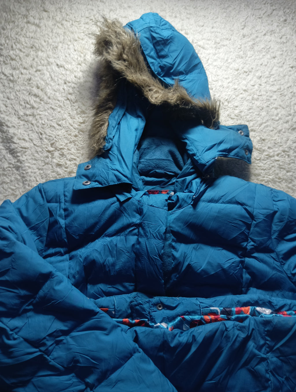 A blue puffer jacket with a fur-trimmed hood, laid flat on a textured white surface. The jacket has horizontal quilting and a patterned fabric detail visible near the waist area.