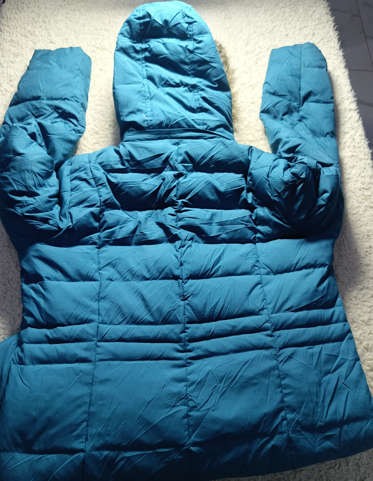 A teal blue puffer jacket with a hood and quilted horizontal stitching, laid flat on a white shag rug. The image has a camera settings overlay at the bottom showing 'POVA 6 Neo' and '23mm f/1.
