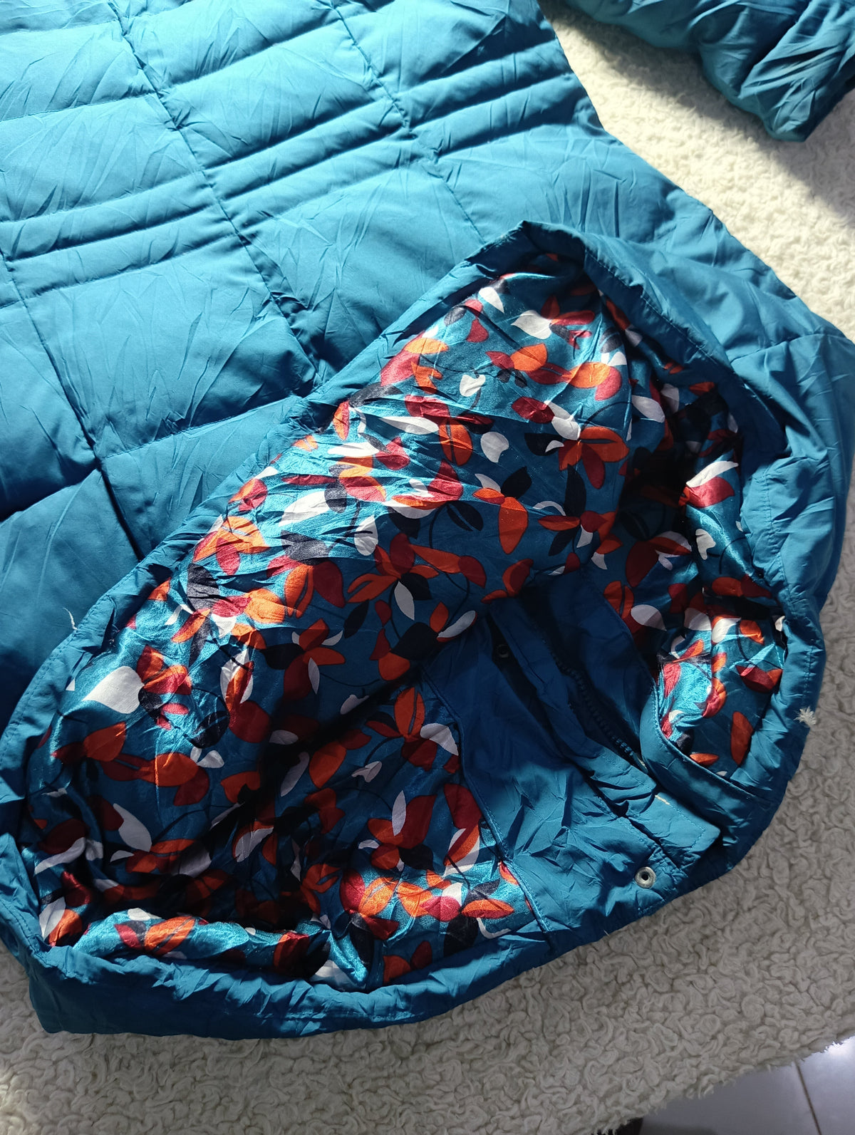 A close-up of a teal blue puffer jacket laid flat on a light textured surface, showing the interior lining with a red, orange, and white floral pattern.