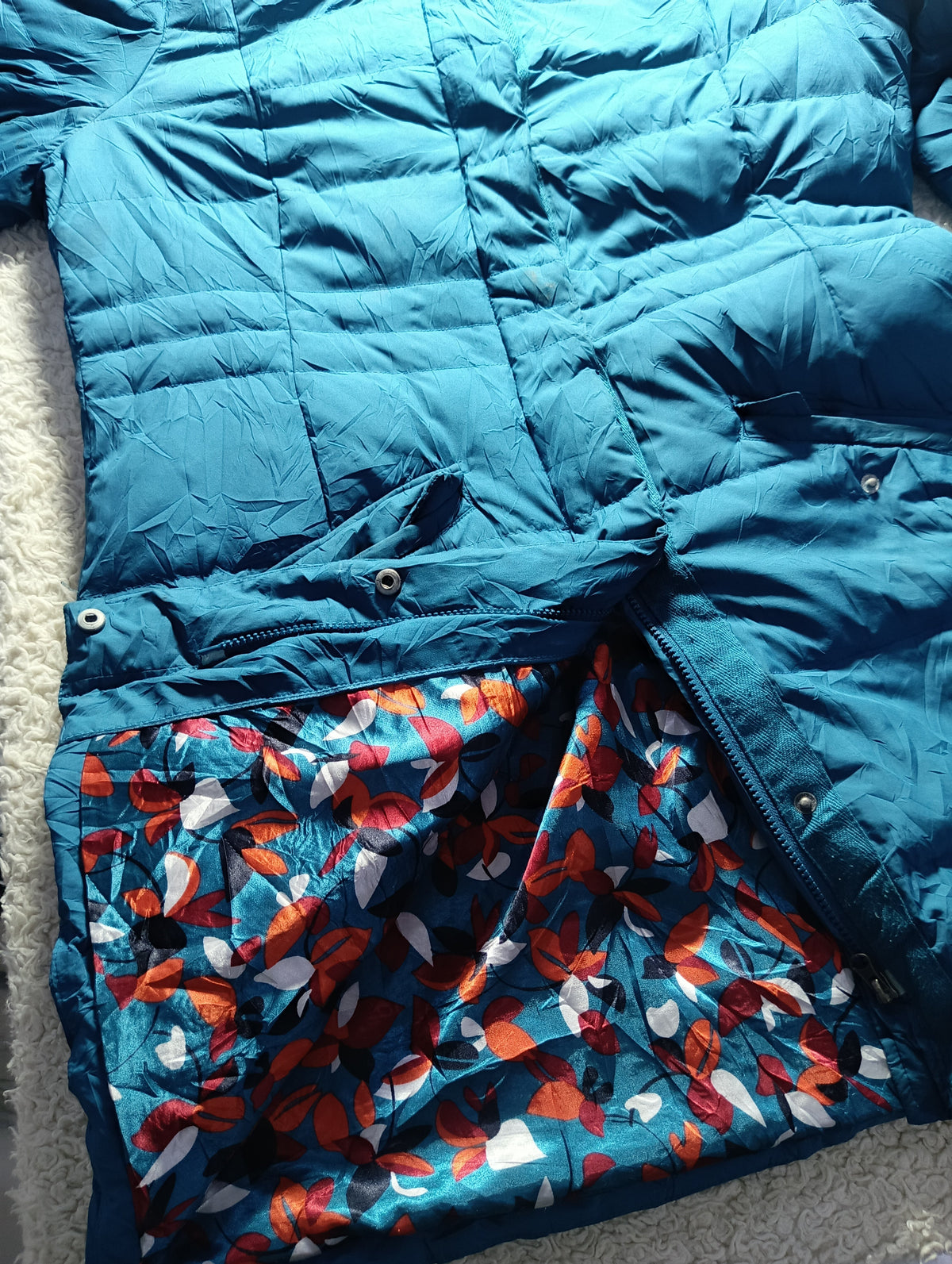 A close-up of a blue puffer jacket laid flat, showing a patterned lining with orange, red, and white leaf-like shapes on a blue background. The photo is taken on a textured light surface with a camera settings watermark at the bottom.