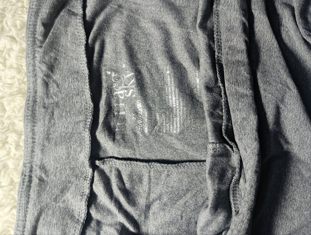 Close-up of a dark gray heathered fabric garment laid flat, showing stitched seams and a faint printed interior label. A light textured surface is visible along the bottom edge.