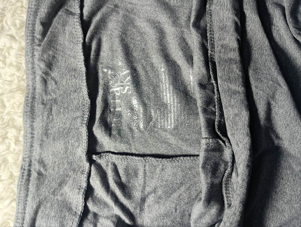 Close-up of a dark gray heathered fabric garment laid flat, showing stitched seams and a faint printed interior label. A light textured surface is visible along the bottom edge.