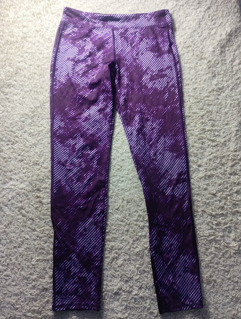 Purple patterned athletic leggings laid flat on a textured gray surface, featuring a diagonal stripe design with a mottled purple and white color scheme.