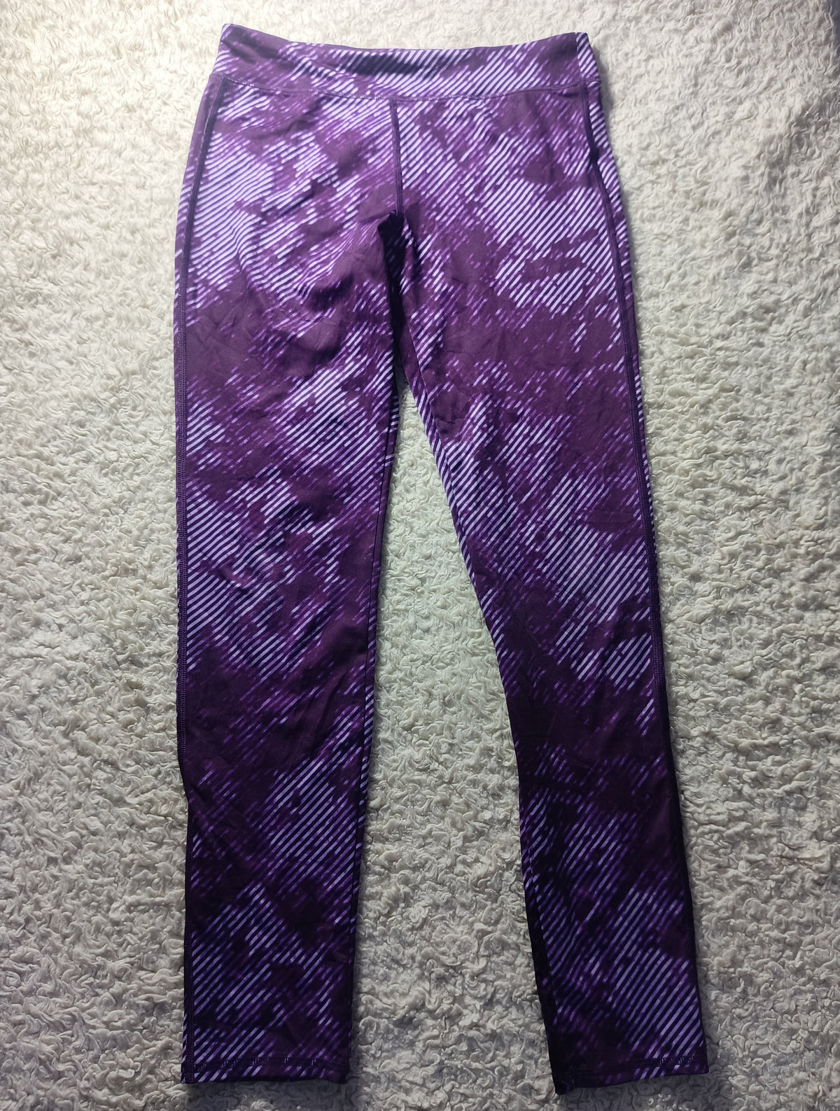 Purple patterned athletic leggings laid flat on a textured gray surface, featuring a diagonal stripe design with a mottled purple and white color scheme.